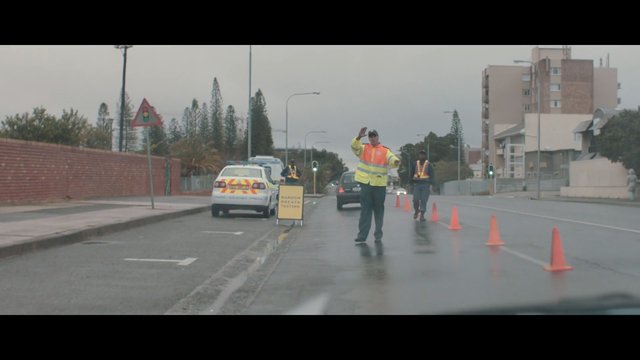 Video Reference: road checkpoint, traffic cones, high visibility vest, police, random breath testing, wet road, urban street, traffic control, vehicle, outdoor, overcast, street scene, safety
