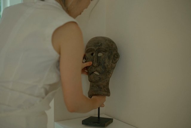 Video Reference: person, sculpture, mask, art, gallery, indoors, installation, stand, white clothing, hand, adjusting, wall, decor, object, closeup