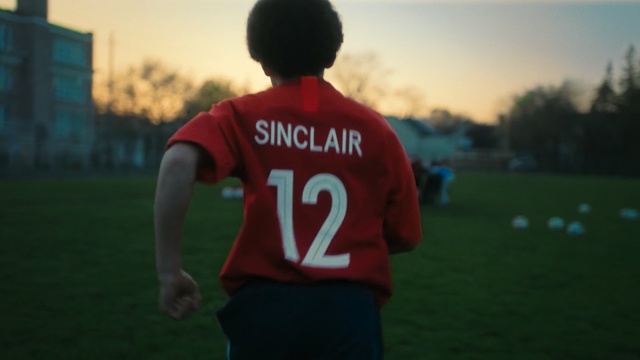 Video Reference: Red, Football player, Football, Green, Player, Youth, Sportswear, Standing, Jersey, Grass, Person, Outdoor, Game, Field, Soccer, Young, Holding, Playing, Ball, Man, Hand, Baseball, Boy, Wearing, Frisbee, Air, White, Flying, Sky, Clothing, Sports uniform, Athletic game