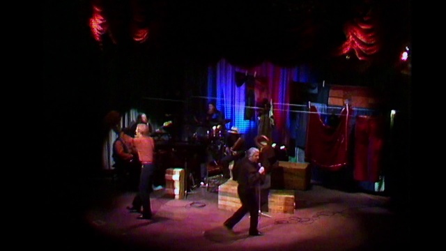 Video Reference: Entertainment, Performance, Stage, Performing arts, Musical theatre, Heater, Performance art, Event, Lighting, Music venue, Outdoor, Street, Light, Dark, Man, Standing, Front, Night, People, Walking, Lit, City, Red, Room, Holding, Woman, Crowd, Group, Concert, Person, Dance, Clothing, Musical instrument, Music