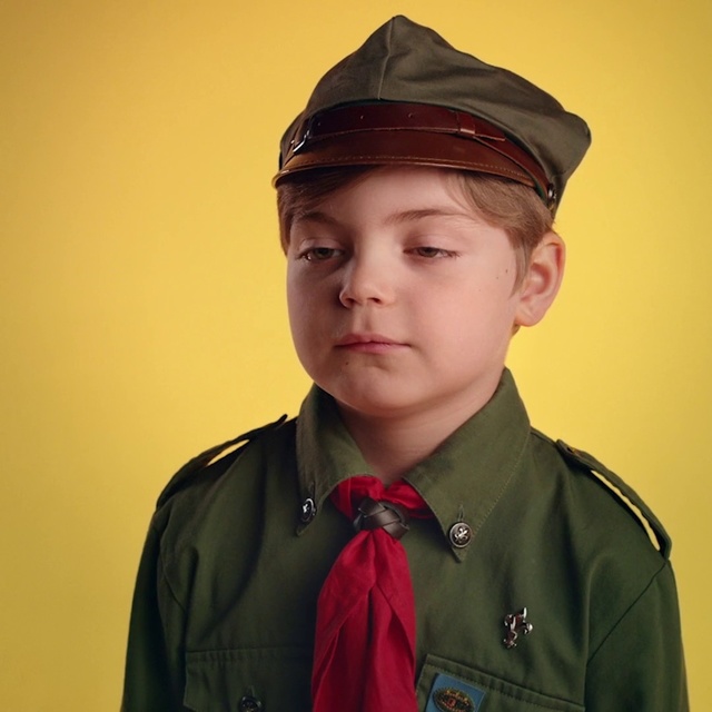 Video Reference: Chin, Cap, Forehead, Headgear, Uniform, Side cap, Neck, Military uniform, Jacket, Fashion accessory, Person, Wearing, Indoor, Hat, Clothing, Man, Boy, Young, Holding, Green, Posing, Dressed, Camera, Standing, Smiling, Suit, Shirt, Baseball, Red, Yellow, Wall, Human face, Peaked cap, Collar