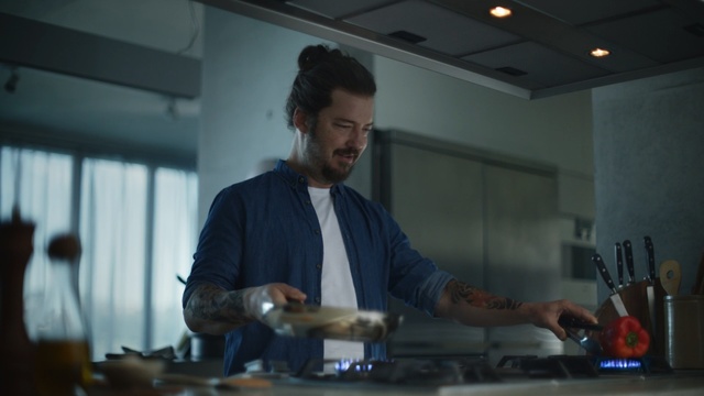Video Reference: Man, Modern kitchen, Kitchen, Cooking, Cook, Smile, Enjoy, Home, Person, Indoor, Ceiling, Preparing, Standing, Food, Table, Holding, Counter, Hand, Cutting, Woman, Shirt, Cake, Pan, Large, Stove, Knife, Cut, Plate, Blue, Room, Oven, People, Pizza, Wall, Clothing, Human face