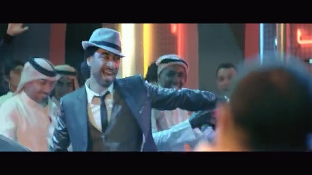 Video Reference: Movie, Gentleman, Fun, Screenshot, Fictional character, Performance, Moustache, Person, Man, Monitor, Photo, Screen, Television, Standing, Suit, Looking, Holding, Food, Table, Glass, Room, Wearing, Large, Display, Bus, Hat, City, Street, People, Phone, Train, Human face, Clothing, Smile, Fashion accessory, Fedora, Text, Dance, Crowd