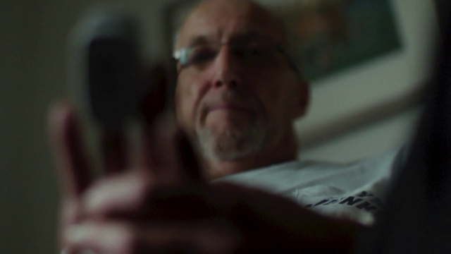 Video Reference: Face, Nose, Head, Skin, Chin, Eyebrow, Facial hair, Mouth, Cheek, Glasses, Person, Indoor, Man, Looking, Sitting, Blurry, Front, Holding, Dark, Room, Television, Close, Laptop, Wearing, Computer, Mirror, Remote, Bed, White, Standing, Human face, Blur, Staring