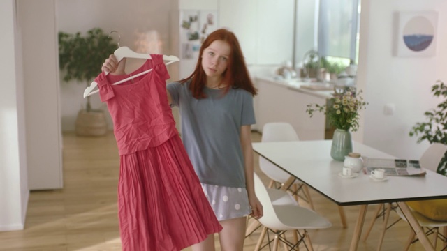 Video Reference: Clothing, Dress, Shoulder, Pink, Gown, Joint, Formal wear, Outerwear, Room, Sleeve, Person, Indoor, Woman, Red, Table, Window, Standing, Looking, Girl, Living, Sitting, White, Dressed, Young, Mirror, Holding, Wearing, Television, Bed, Cat, Shower, Playing, Floor, Wall, Skirt