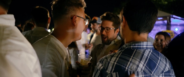 Video Reference: Event, Eyewear, Fun, Design, Glasses, Crowd, Vision care, Conversation, Person, Man, Indoor, Wine, Looking, People, Table, Standing, Glass, Drinking, Woman, Holding, Group, Food, Couple, Young, Plate, Red, Phone, Room, Pizza, Human face, Clothing, Drink