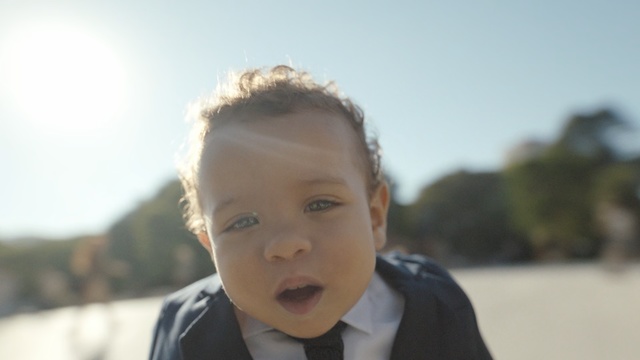 Video Reference: Face, Child, Hair, Facial expression, People, Head, Nose, Cheek, Forehead, Male, Person, Outdoor, Young, Looking, Wearing, Woman, White, Boy, Front, Man, Sitting, Shirt, Suit, Smiling, Standing, Eating, Holding, Sky, Toddler, Baby, Human face, Clothing