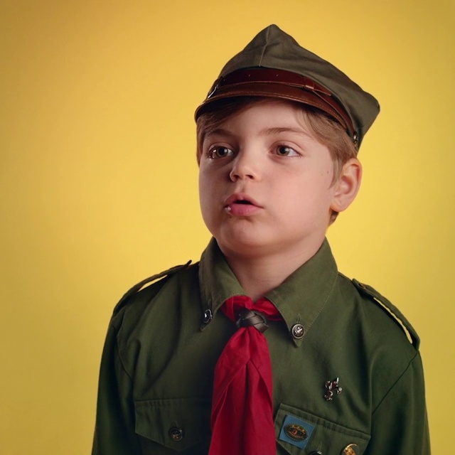 Video Reference: Chin, Cap, Headgear, Uniform, Side cap, Hat, Military uniform, Costume hat, Portrait, Sleeve, Person, Wearing, Green, Clothing, Indoor, Necktie, Man, Jacket, Boy, Young, Standing, Posing, Dressed, Suit, Holding, Shirt, Camera, Front, Smiling, Hair, Red, Bow, Yellow, Umbrella, White, Wall, Fashion accessory, Human face, Fedora, Collar, Toddler, Coat