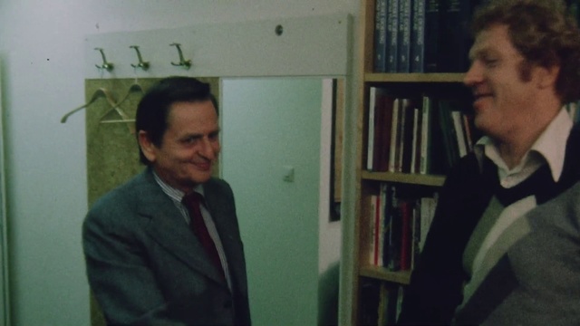 Video Reference: Conversation, Professor, White-collar worker, Suit, Official, Person, Man, Indoor, Necktie, Standing, Looking, Front, Holding, Wearing, Book, Mirror, Room, Green, Young, Table, White, Shirt, Computer, Wall, Bookcase, Shelf, Text, Human face