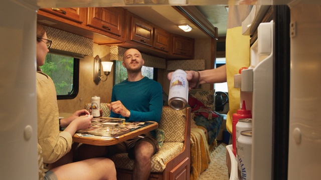 Video Reference: Room, Interior design, Vacation, Table, Rv, Leisure, Furniture, Indoor, Person, Cabinet, Kitchen, Small, Man, Sitting, Holding, Refrigerator, Filled, Woman, Sink, Standing, Young, White, Living, Blue, Bottle, Clothing, Smile, Chair, Drink, Human face, Food