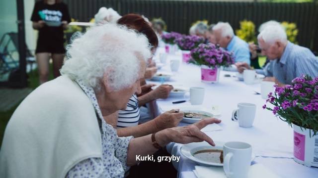 Video Reference: Grandparent, Event, Lunch, Meal, Plant, Flower, Floral design, Tableware, Person, Table, Food, Woman, People, Man, Looking, Standing, Sitting, Front, Cake, Eating, Group, Phone, Older, Cutting, Fruit, Plate, Sandwich, Holding, Laptop, Bowl, Crowd, White, Sheep, Wedding, Kitchen, Vase, Coffee cup