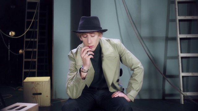Video Reference: Clothing, Suit, Gentleman, Hat, Male, Fedora, Fashion, Headgear, Outerwear, Fashion accessory, Person, Man, Wearing, Sitting, Holding, Cellphone, Phone, Table, Dressed, Standing, Woman, Talking, Black, Using, Young, Computer, Laptop, White, Sun hat, Cowboy hat, Human face, Coat, Costume hat