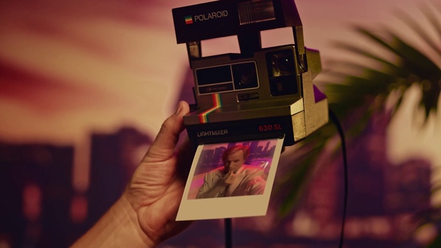 Video Reference: Cameras & optics, Instant camera, Pink, Photography, Hand, Camera, Magenta, Room, Film camera, Camera accessory, Indoor, Sitting, Holding, Table, Small, Front, Man, Laptop, Phone, Screen, Computer, Display, Keyboard, Standing, Book, Text, Screenshot