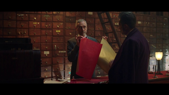 Video Reference: Screenshot, Adaptation, Scene, Event, Person, Man, Table, Standing, Red, Holding, Front, Woman, Orange, Suit, Wearing, Computer, Phone, Food, Laptop, Large, Display, Room, White, People, Human face, Clothing