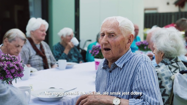 Video Reference: Community, Grandparent, Event, Adaptation, Retirement home, Lunch, Person, Man, Table, Woman, People, Cake, Standing, Older, Front, Holding, Food, Group, Wearing, Walking, Street, Talking, White, Large, Birthday, Cutting, Blue, Crowd, Plate, Room, Human face, Clothing, Senior citizen