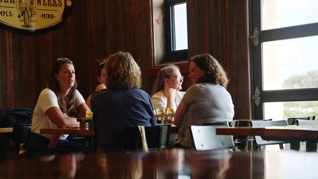 Video Reference: Event, Conversation, Interaction, Room, Meeting, Restaurant, Drink, Person, Sitting, Table, Indoor, Window, People, Woman, Looking, Group, Front, Food, Man, Wooden, Eating, Computer, Standing, Young, Clothing, Human face, Bottle, Meal