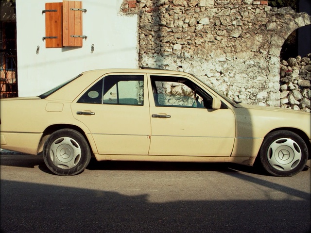 Video Reference: Land vehicle, Vehicle, Car, Mercedes-benz w124, Alloy wheel, Luxury vehicle, Mercedes-benz, Classic car, Rim, Mercedes-benz w201, Building, Road, Outdoor, Parked, Front, Street, Truck, House, Sitting, Old, Small, Side, Driving, Yellow, City, Parking, Brick, Traffic, Stop, Man, Riding, Sign, White, Standing, Bed, Wheel, Auto part, Curb, Roof