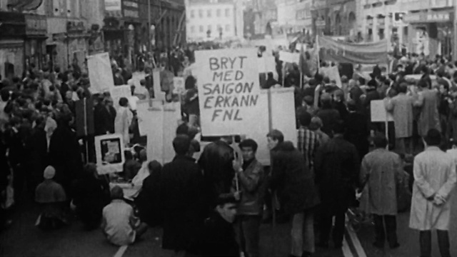 Video Reference: Crowd, Black-and-white, Event, Monochrome, Rebellion, Monochrome photography, Street, History, City, Person, Building, People, Photo, White, Group, Walking, Black, Many, Crowded, Old, Busy, Standing, Large, Filled, Sign, Luggage, Umbrella, Train, Room, Man, Holding, Suitcase, Station, Clothing, Black and white, Presentation, Text, Handwriting, Woman