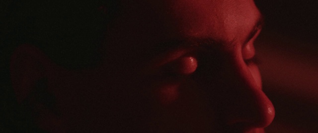 Video Reference: Red, Face, Black, Nose, Lip, Darkness, Head, Ear, Light, Mouth, Person, Man, Indoor, Looking, Holding, Dark, Wearing, Front, Sitting, Glasses, Close, Room, Food, Teeth, Orange, Laptop, Computer, Shirt, Young, Yellow, Standing, Brushing, White, Staring, Blur