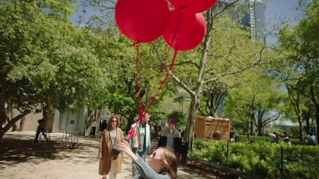 Video Reference: Red, Balloon, Tree, Party supply, Organism, Plant, Leisure, Adaptation, Architecture, Flower, Outdoor, Walking, Standing, Woman, Street, Holding, Young, Park, Man, Large, Wearing, Boy, Girl, Umbrella, Bear, Hat, Riding, Playing, People, Clothing, Outdoor object