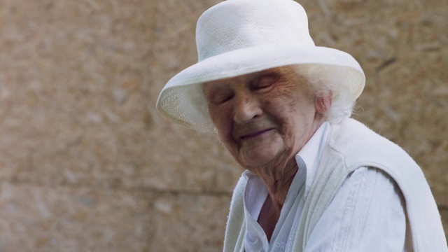 Video Reference: Skin, Hat, Wrinkle, Headgear, Human, Elder, Smile, Gentleman, Cowboy hat, Person, Building, Man, Wearing, Standing, Looking, Holding, White, Front, Head, Suit, Red, Large, Phone, Fashion accessory, Human face, Sun hat, Fedora, Clothing