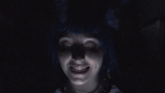 Video Reference: Face, Black, Facial expression, Head, Smile, Darkness, Nose, Eye, Fiction, Mouth, Person, Clothing, Woman, Looking, Young, Posing, Holding, Wearing, Smiling, Dark, Camera, Standing, Front, Dress, Girl, White, Red, Shirt, Man, Room, Hat, Blue, Human face, Portrait