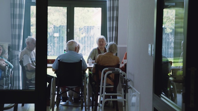 Video Reference: Window, Retirement home, Room, Wheelchair, Glass, Restaurant, Person, Indoor, Table, Chair, Sitting, Building, Man, Woman, Front, Food, People, Living, Dining, Wine, White, Young, Laptop, Group, Eating, Large, Kitchen, Standing, Computer, Dog, City, Phone, Furniture, Clothing, Dining table