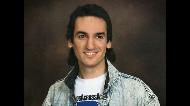 Video Reference: Hair, Facial expression, Eyebrow, Chin, Forehead, Cheek, Smile, Hairstyle, Jaw, Portrait, Person, Man, Indoor, Clothing, Necktie, Front, Shirt, Wearing, Smiling, Looking, Posing, Photo, Holding, Suit, Young, Camera, Dark, Table, Sitting, White, Laptop, Standing, Glasses, Large, Woman, Human face, Wall, Face, Headshot, Text, Male