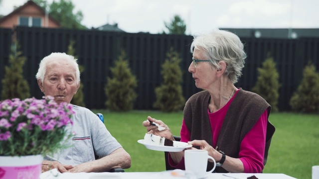 Video Reference: Community, Grandparent, Event, Fun, Grass, Plant, Recreation, Leisure, Flower, Person, Outdoor, Woman, Table, Sitting, Food, Plate, People, Older, Cake, Holding, Park, Bench, Pink, Glass, Standing, Man, White, Group, Clothing, Human face, Glasses