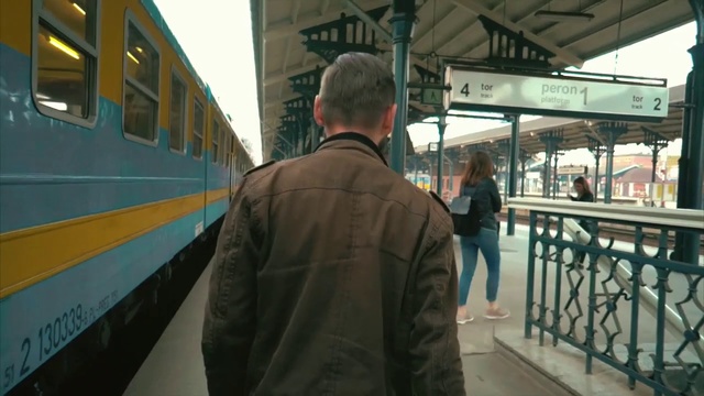 Video Reference: Transport, Public transport, Rolling stock, Train station, Train, Passenger, Mode of transport, Metro, Vehicle, Passenger car, Person, Outdoor, Building, Man, Bus, Standing, Station, Platform, Green, Yellow, Walking, Waiting, Holding, Sidewalk, Subway, People, Woman, Blue, Red, Track, Board, Rain, Parked, Luggage, Clothing, Land vehicle, Text