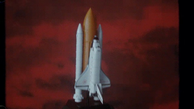 Video Reference: Space shuttle, Rocket, Missile, Spacecraft, Spaceplane, Vehicle, Space, Rocket-powered aircraft, Aircraft, Indoor, Sitting, Dark, Front, Close, Table, Red, Light, Blurry, Wooden, Room, Computer, White, Engine, Airplane, Aerospace engineering, Aerospace manufacturer, Aviation, Airliner, Air travel, Transport, General aviation, Airline, Jet aircraft, Monoplane, Flap, Air racing, Supersonic aircraft, Flight, Air