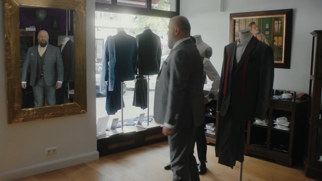 Video Reference: Suit, Room, Event, Formal wear, Collection, Person, Indoor, Man, Standing, Front, Window, Holding, Wearing, Group, Table, Mirror, Woman, Television, Large, Living, White, People, Floor, Clothing, Wall, Coat, Trousers, Footwear, Outerwear