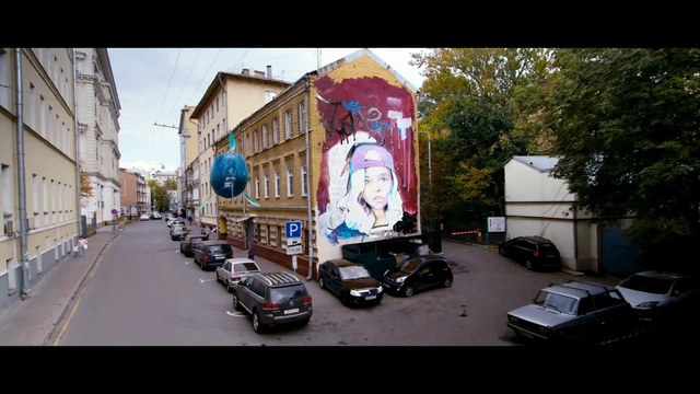 Video Reference: Neighbourhood, Town, Street art, Mode of transport, Street, Mural, Advertising, Transport, Art, Urban area, Person, Outdoor, Road, Building, Car, Riding, Traffic, Man, City, Side, Table, Red, Light, Parked, Stop, Driving, Standing, People, Motorcycle, Text, Land vehicle, Vehicle, Billboard, Way