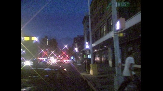 Video Reference: Street light, Metropolitan area, Night, Lighting, Light, Urban area, Metropolis, Sky, Darkness, Light fixture, Outdoor, Building, Street, Traffic, City, Dark, Busy, Sitting, Stop, Road, Intersection, Lit, Sign, Driving, Red, Standing, People, Signal, Blurry, Text, Skyscraper