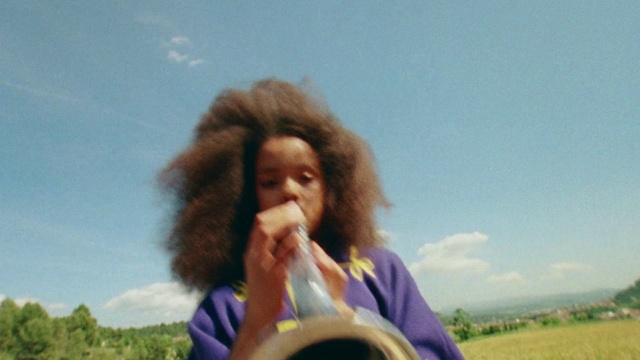 Video Reference: Musical instrument, Fun, Afro, Wind instrument, Happy, Woodwind instrument, Person, Outdoor, Grass, Man, Woman, Holding, Eating, Field, Standing, Young, Shirt, Wearing, Green, Water, Grassy, Playing, White, Sky, Human face, Clothing, Nature, Girl
