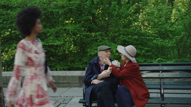 Video Reference: Botany, Sitting, Adaptation, Event, Leisure, Person, Outdoor, Bench, Park, Man, Child, Young, Woman, Boy, People, Holding, Game, Group, Red, Phone, Tree, Hat, Clothing, Fashion accessory, Sun hat, Fedora, Cowboy hat