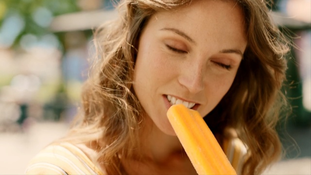 Video Reference: Junk food, Eating, Lip, Food craving, Food, Long hair, Brown hair, Smile, Ice pop, Person, Woman, Outdoor, Girl, Lady, Mouth, Holding, Orange, Street, Young, Female, Sitting, Yellow, Wearing, Standing, Pink, White, Fast food, Human face, Face, Beautiful, Dessert