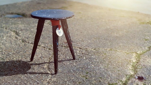 Video Reference: stool, tripod stand, wooden legs, wet pavement, sunlight, outdoor, close-up, small object, concrete, shadow, tag, handmade, rustic, minimal scene