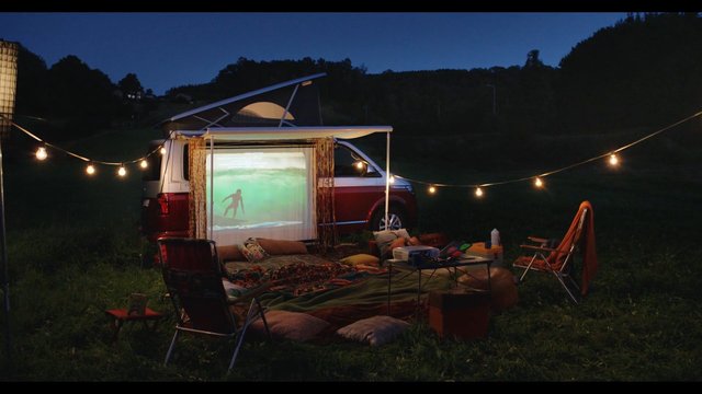 Video Reference: campsite, night, van, projector, outdoor movie, string lights, folding chairs, blankets, grass, surfer, screen, camping, recreation, travel, evening, hills