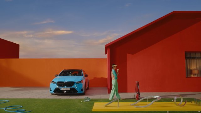 Video Reference: blue car, red building, orange wall, person, ice cream, courtyard, modern, colorful, outdoor, minimalist, architecture, parked car, sky, grass, bright, stylized