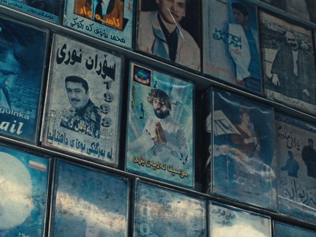 Video Reference: posters, framed pictures, wall art, music posters, portraits, Arabic text, Persian text, indoor wall, tilted view, collage, vintage, blue tones, media display
