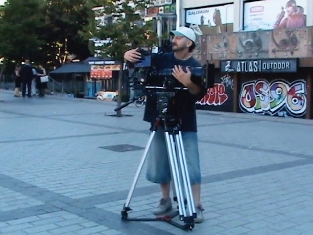 Video Reference: man, video camera, tripod, filming, outdoor plaza, graffiti, street scene, urban, cameraman, standing, public space, building facade, people in background
