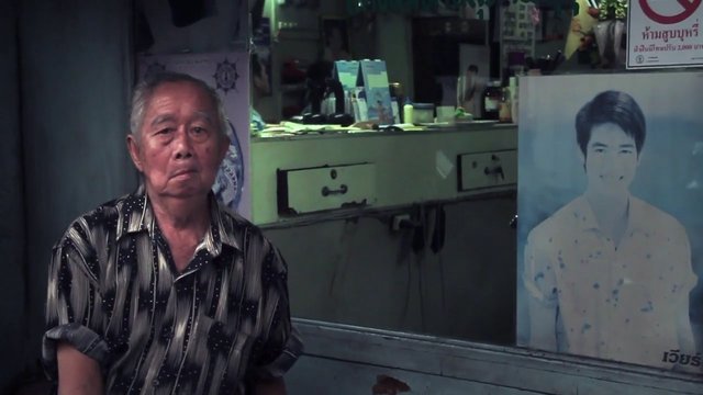 Video Reference: elderly man, indoor, portrait poster, Thai text, shop interior, counter, window display, seated, poster, sign, reflection, interview scene