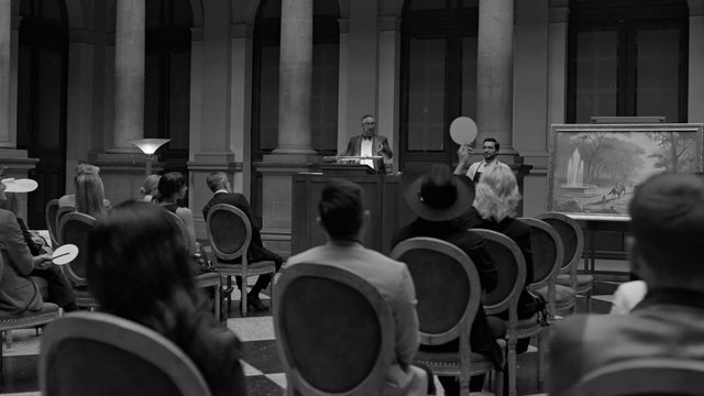 Video Reference: auction, indoor, audience, speaker, podium, painting, art sale, historic building, black and white, event, seated crowd, formal attire, bid paddle, gallery, columns