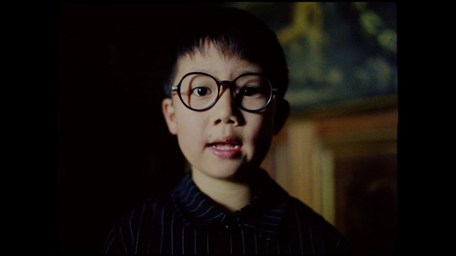 Video Reference: child, portrait, glasses, close-up, indoor, dim light, face, speaking, young person, dark background, shallow depth of field