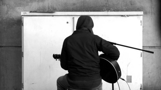 Video Reference: person, guitar, musician, back view, indoors, concrete wall, black and white, seated, instrument, playing guitar, cabinet, door, hooded jacket, practice, music