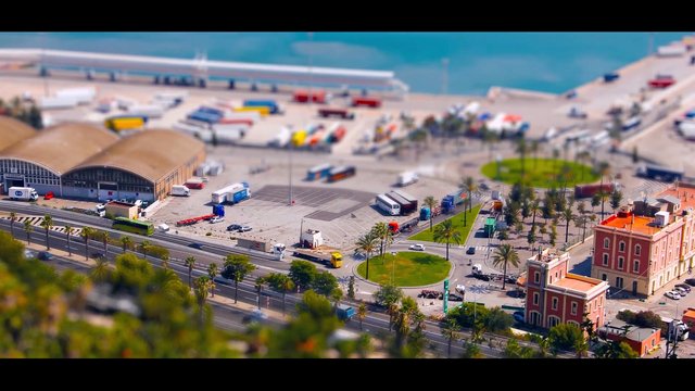 Video Reference: port, harbor, coastal, aerial, high-angle, trucks, buses, road, palm trees, buildings, waterfront, tilt-shift, industrial, cityscape, transport, parking