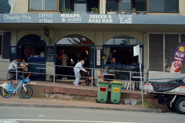 Video Reference: cafe, storefront, outdoor seating, street scene, people, bicycle, delivery truck, menu sign, suburban, sidewalk, restaurant, outdoor dining, daytime, urban, building facade, acai bar