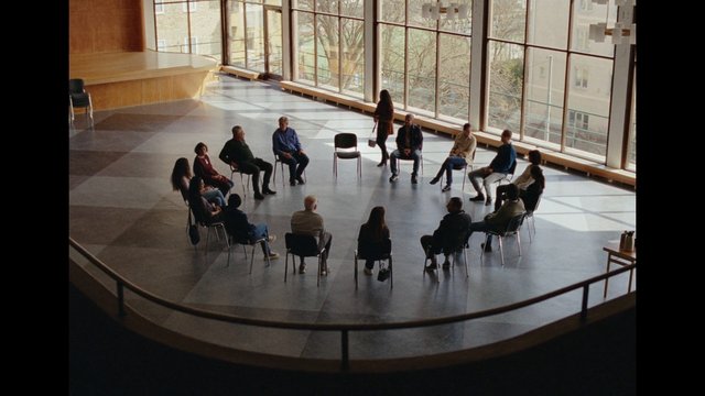 Video Reference: people, group meeting, circle, chairs, indoor, sunlit, windows, large room, discussion, waiting room, auditorium, interior, standing person, community, wide shot