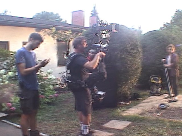 Video Reference: backyard, garden, people, camera, filming, crew, outdoors, house, hedge, tripod, equipment, casual, daylight, residential, setup
